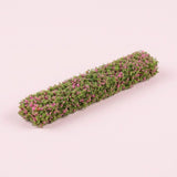 A model hedge with green foliage and small pink floral details rests diagonally on a pale pink background.