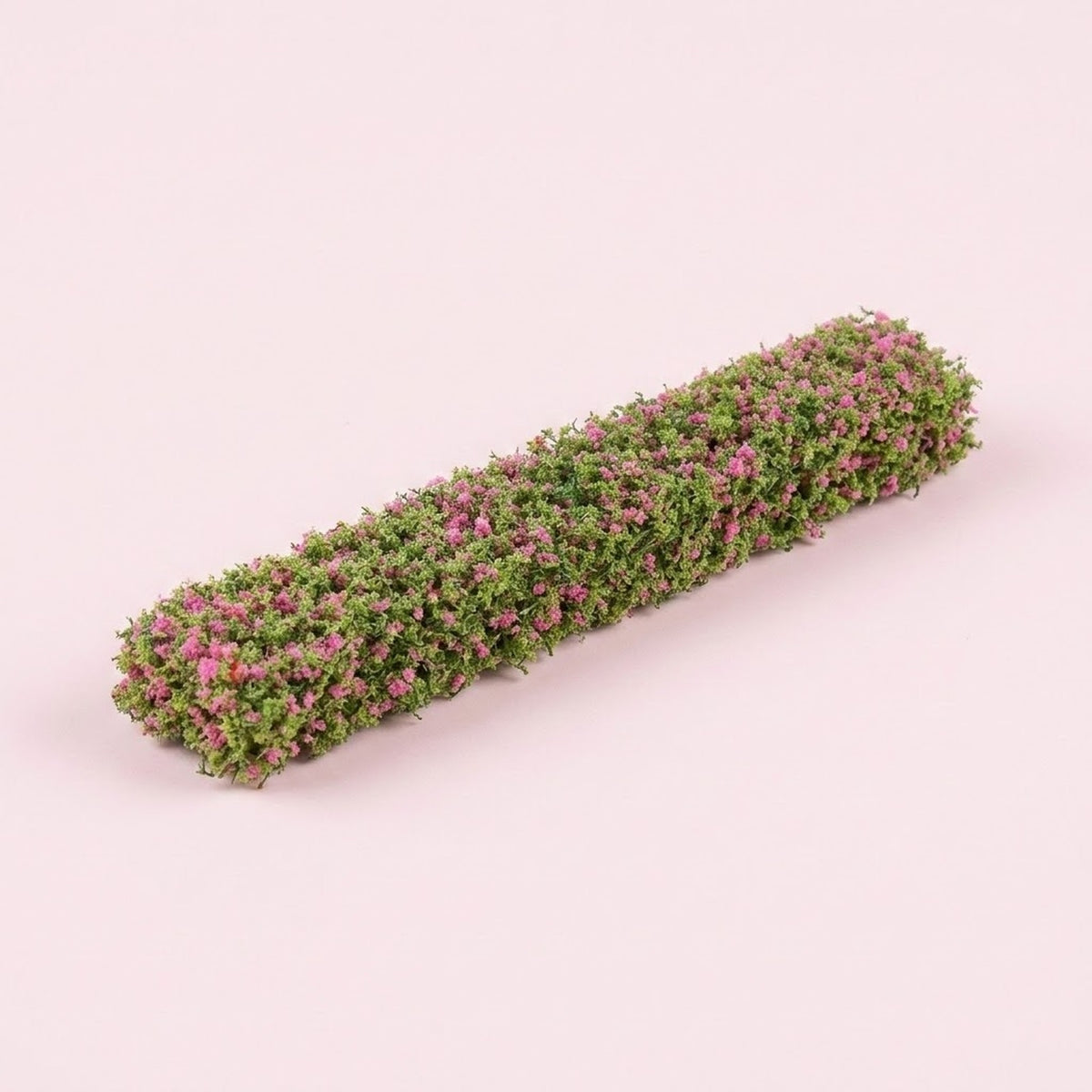 A model hedge with green foliage and small pink floral details rests diagonally on a pale pink background.