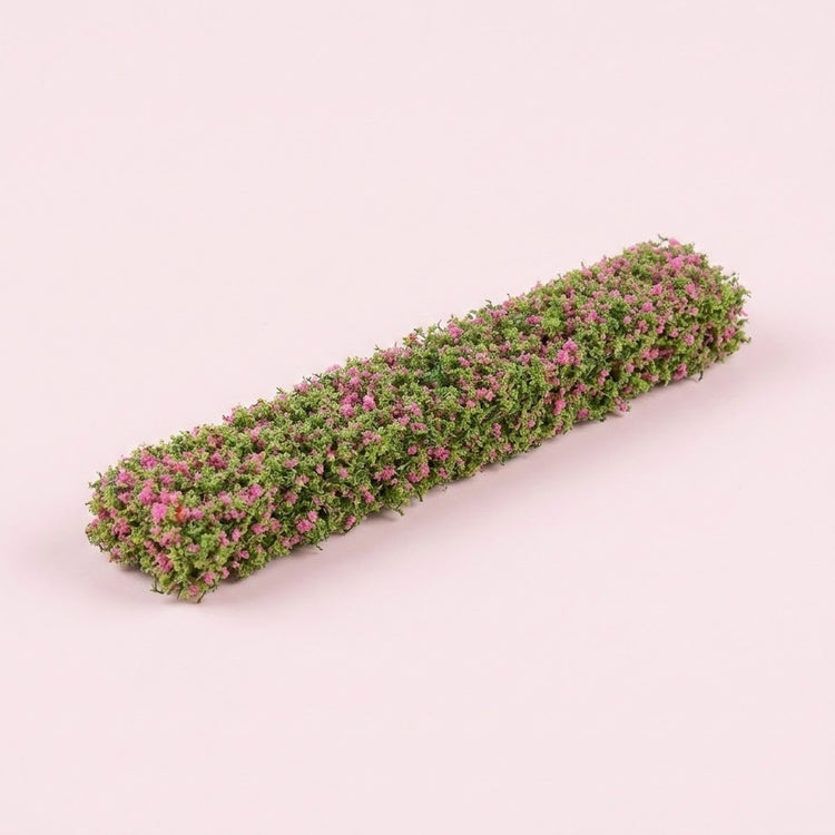 A model hedge with green foliage and small pink floral details rests diagonally on a pale pink background.