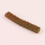 A miniature model hedge with red and green textured foliage lies diagonally on a pale pink background.
