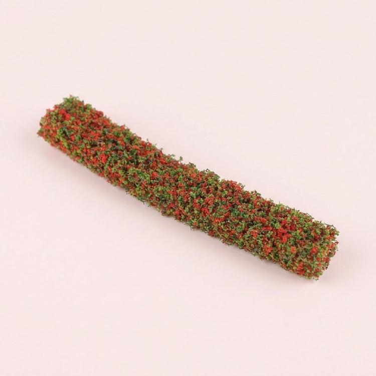 A miniature model hedge with red and green textured foliage lies diagonally on a pale pink background.