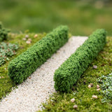 Two green model hedges border a pale gravel path surrounded by miniature grass, plants, and scattered stones.