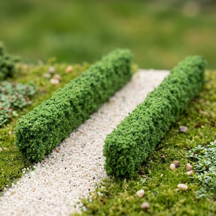 Two green model hedges border a pale gravel path surrounded by miniature grass, plants, and scattered stones.