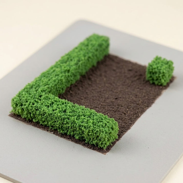 Green miniature hedges form an L-shaped border around brown model terrain, with a separate short shrub at the opposite corner.