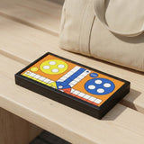 A folded ludo set with a black frame and colourful orange board rests on a light wood bench beside a beige bag.