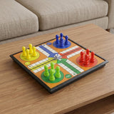 Black-framed Ludo board with red, yellow, green and blue pawns and a white die on a wooden table beside a beige sofa.