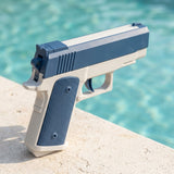Blue-and-white water squirt gun with a textured grip stands on pale poolside stone, viewed from behind against turquoise water.
