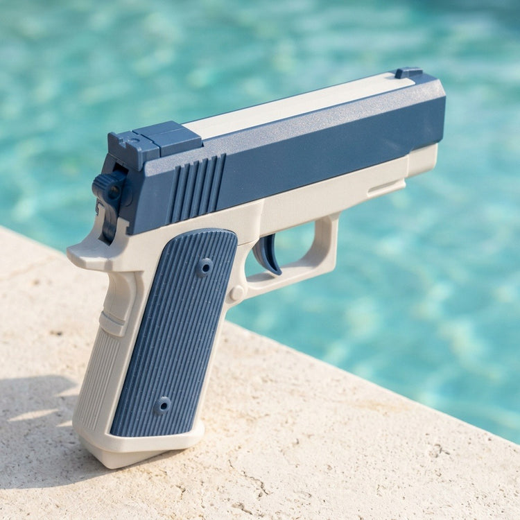 Blue-and-white water squirt gun with a textured grip stands on pale poolside stone, viewed from behind against turquoise water.