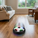 Illuminated black-and-white hover soccer ball with a motion trail on a wooden living-room floor between a cream sofa and brown armchairs.