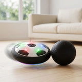 Illuminated black-and-white hover soccer ball beside a round black ball on a pale wooden floor, with a cream sofa behind.