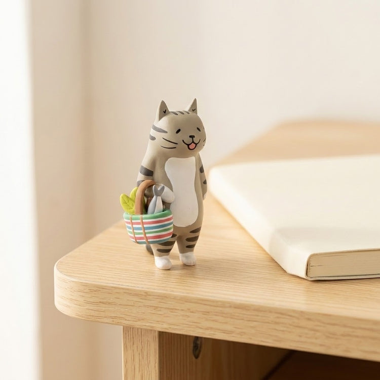 A gray tabby cat blind box figure holds a striped basket on a wooden table beside an open book.