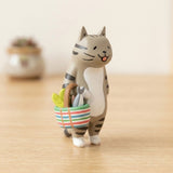 A gray tabby cat blind box figure carries fish and greens in a striped basket on wood against a cream background.