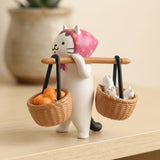 White cat blind box figure wears a pink headscarf and carries two baskets on a shoulder pole, standing on a wooden surface.