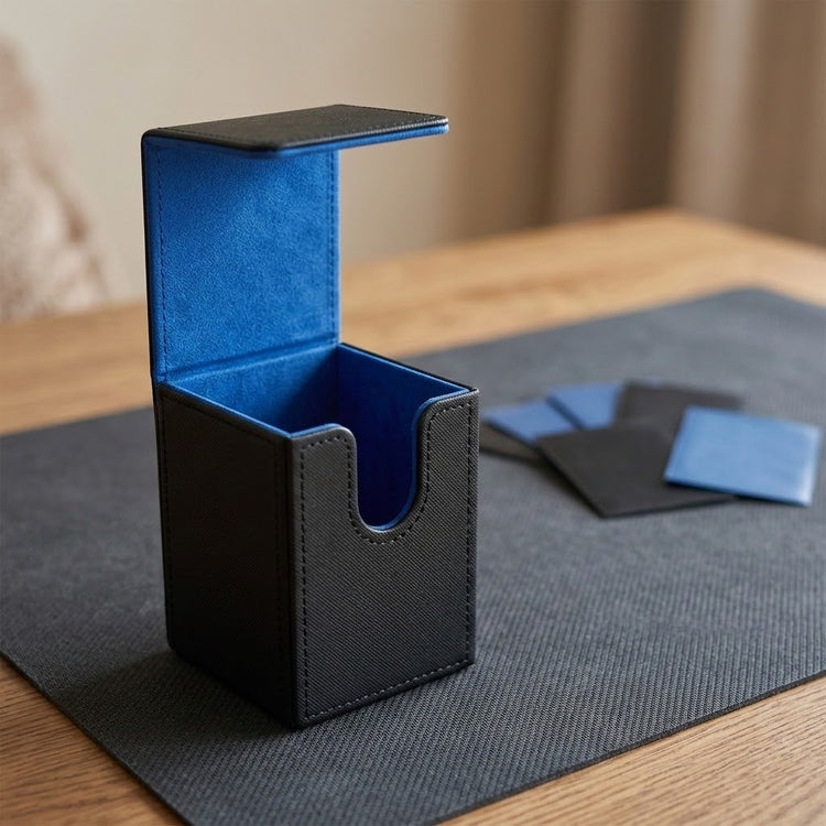 A black textured card holder or deck box with a bright blue velvet interior, shown open on a dark gray mat over a wooden table. A few matching black and blue cards or inserts are placed nearby.