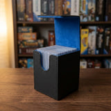 Open black deck box with blue lining holding sleeved cards on a wooden table, with blurred game shelves behind it.