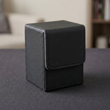 A closed black textured deck box with a stitched front flap stands on a dark gray tabletop in a softly blurred room.