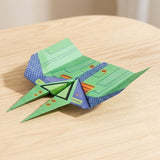 Green and blue pre-printed paper airplane from this origami kit, with broad patterned wings and two pointed tips, rests on a light wooden table.