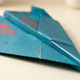 Close-up of a teal-blue paper airplane from this origami kit with red printed accents, resting on a light beige surface.