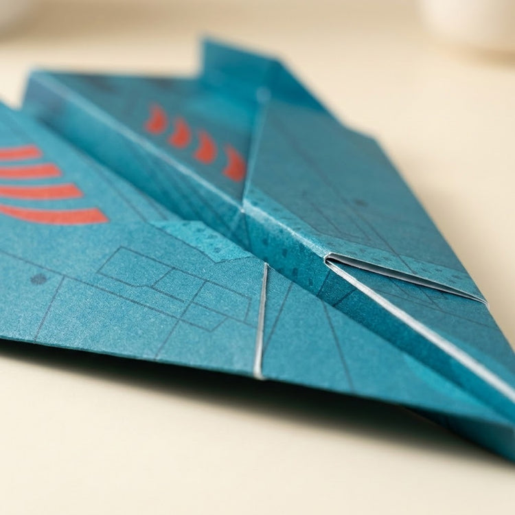 Close-up of a teal-blue paper airplane from this origami kit with red printed accents, resting on a light beige surface.