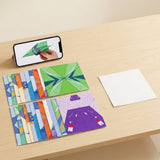 Colorful printed origami kit sheets and a blank white sheet sit on a wooden table beside a smartphone showing paper airplane folding.