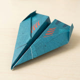 Teal paper airplane folded from this origami kit with orange wing markings and upturned edges, resting on a pale wooden surface.