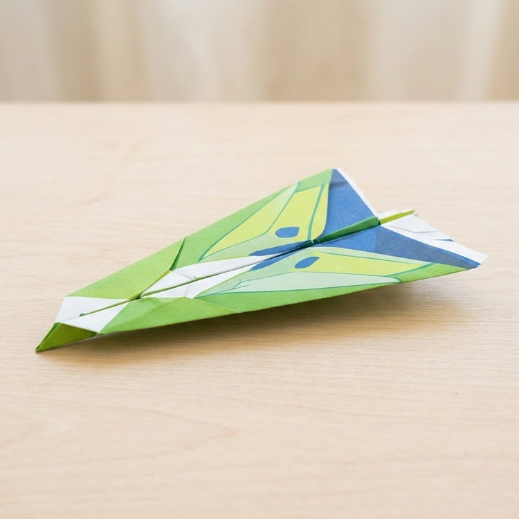 Green, yellow, blue, and white paper airplane folded from this origami kit on a light wooden table against a beige background.