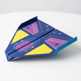 A dark blue paper airplane from the origami kit has yellow and purple graphics and upturned wingtips against a white background.