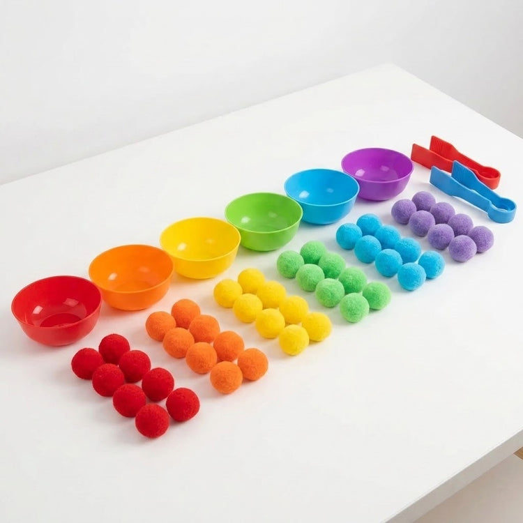 Sorting toy with six rainbow plastic bowls beside color-grouped pom poms and red and blue tweezers on a white table.