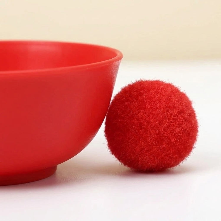 Sorting toy detail showing a red plastic bowl beside a fluffy red pom pom on a white surface against a beige background.