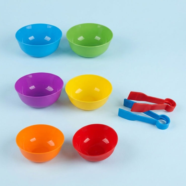 Sorting toy accessories on a light blue background: six empty plastic bowls in rainbow colors with red and blue tweezers.