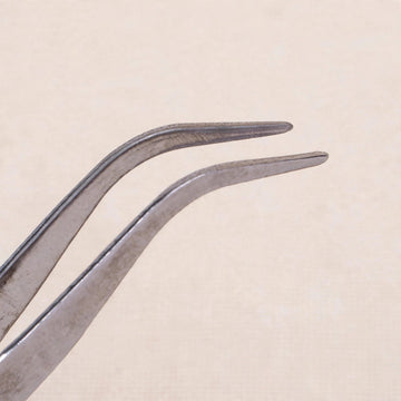 Silver metal tweezers with bent tips shown close up against a pale beige background.