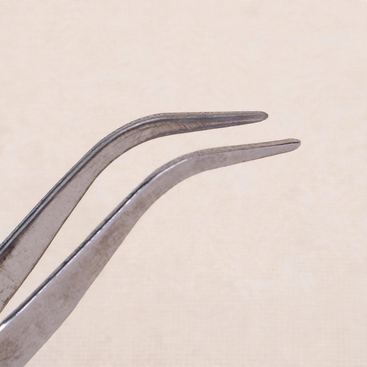 Silver metal tweezers with bent tips shown close up against a pale beige background.