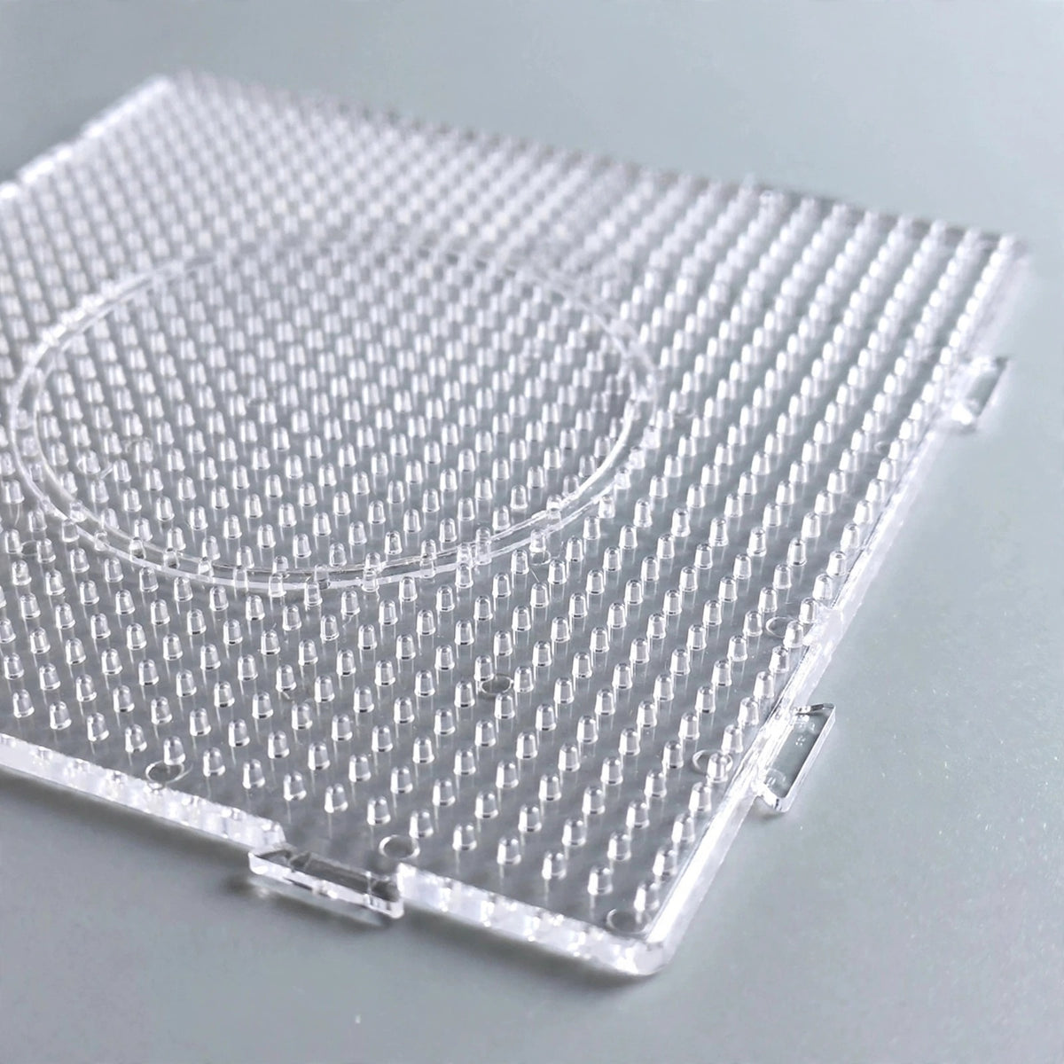 A clear square plastic bead pegboard with a circular outline and interlocking edges rests on a grey surface.