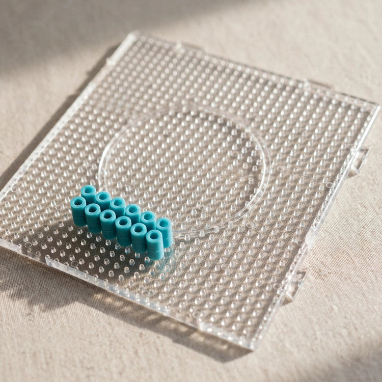 Turquoise cylindrical beads form two short rows on a clear square pegboard resting on beige fabric.