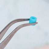 Bent silver metal tweezers hold a single turquoise cylindrical bead against a pale background.