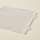 A clear square plastic bead pegboard with rows of upright pegs and interlocking tabs rests on a cream surface.