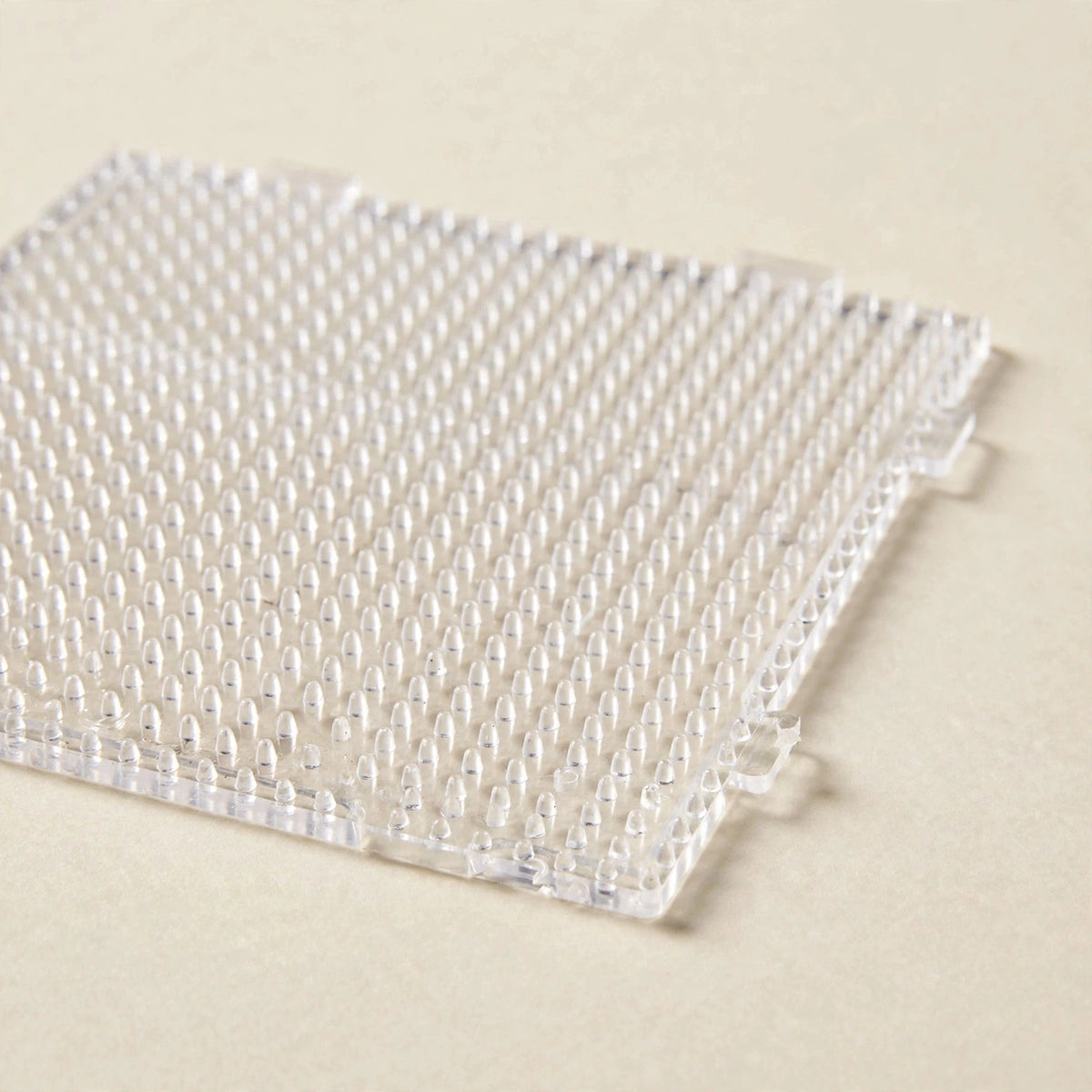 A clear square plastic bead pegboard with rows of upright pegs and interlocking tabs rests on a cream surface.