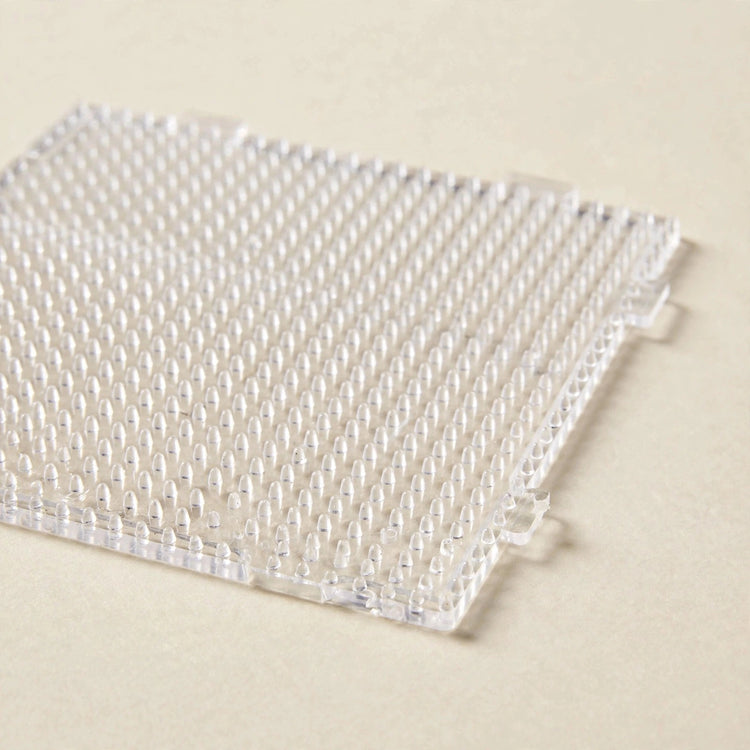 A clear square plastic bead pegboard with rows of upright pegs and interlocking tabs rests on a cream surface.