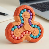 A three-spoke fidget puzzle toy with an orange base and colorful inner beads, shown on a light desk with a blurred laptop in the background.