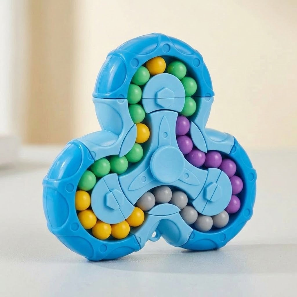 A blue three-spoke fidget puzzle toy filled with small round beads in green, yellow, purple, and gray, photographed on a light neutral surface with a softly blurred background.
