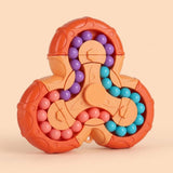 A three-spoke fidget puzzle toy with orange plastic outer rings, peach-colored center pieces, and rows of small round beads in purple, turquoise, and pink, shown on a warm beige background.