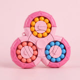 A pink plastic fidget puzzle displays three circular tracks of yellow, coral and blue beads against a pale pink background.