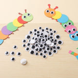 A craft activity set of googly eyes and white pegs displayed on a light wood surface, with two colorful paper caterpillar models demonstrating how to assemble the craft.