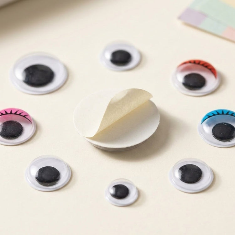 Black and multicolor plastic googly eyes surround an overturned eye with partially peeled backing on a cream surface.