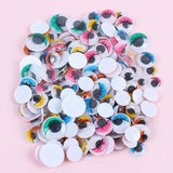 Multicolor plastic googly eyes with black pupils, eyelash details, and white backs form a pile on a pale pink background.