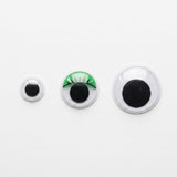Three plastic googly eyes in increasing sizes sit on a white background, with green eyelash detailing on the middle eye.