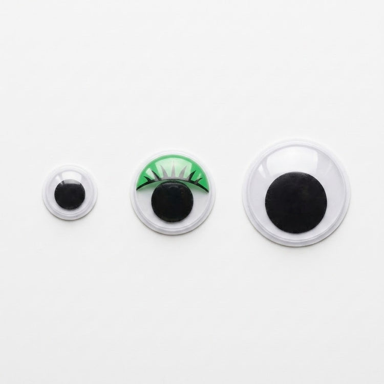 Three plastic googly eyes in increasing sizes sit on a white background, with green eyelash detailing on the middle eye.