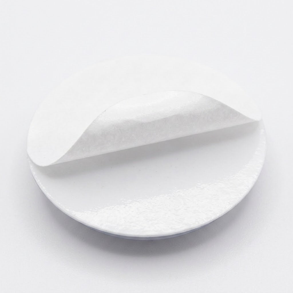 A plastic googly eye rests backing-side up with its white adhesive liner partially peeled away against a white background.