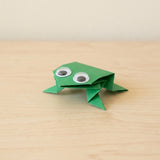 A small green origami frog with googly eyes sits on a light wood tabletop against a soft beige wall.