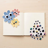 A set of handmade craft decorations displayed on an open book, including three colorful paper flowers with googly eyes on the left page and a pile of assorted googly eyes arranged in a tree shape on the right page, all on a light wood background.